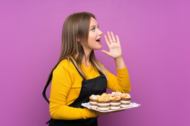 Das Jugendlichmädchen, das viele verschiedene Minikuchen purpurrot schreit mit dem breiten Mund hält, öffnen sich