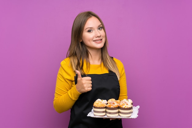 Das Jugendlichmädchen, das viele verschiedene Minikuchen über dem lokalisierten purpurroten Geben Daumen hält, up Geste