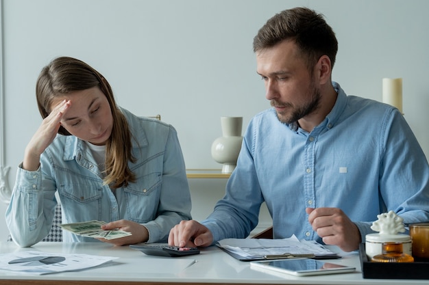 Das junge ehepaar, das am wohnzimmertisch sitzt und papiere studiert, berechnet das familienbudget auf einem taschenrechner