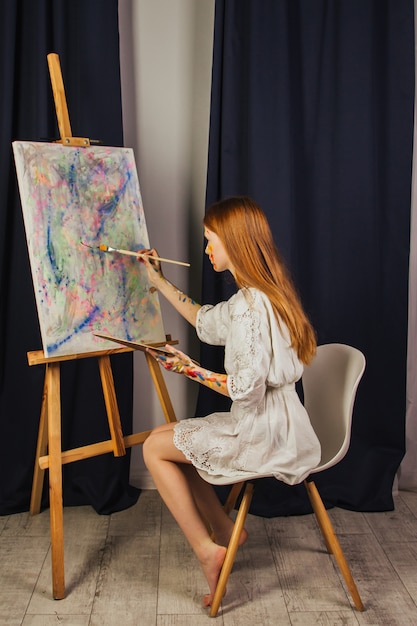 Das junge Mädchen des Künstlers in einem hellen weißen Kleid malt in der Werkstatt ein Bild auf Leinwand. Das Gesicht ist mit Farben befleckt. Ein junger Student benutzt Pinsel, Leinwände und Staffeleien. Kreative Arbeit.