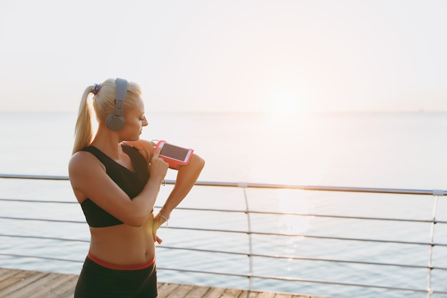 Das junge schöne athletische Mädchen mit langen blonden Haaren in Kopfhörern betrachtet das Handy bei Sonnenaufgang über dem Meer mobile