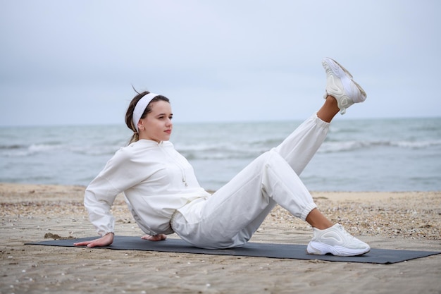 Das junge schöne Mädchen, das auf der Yogamatte liegt und ihr Bein am Strand hochhebt Foto in hoher Qualität