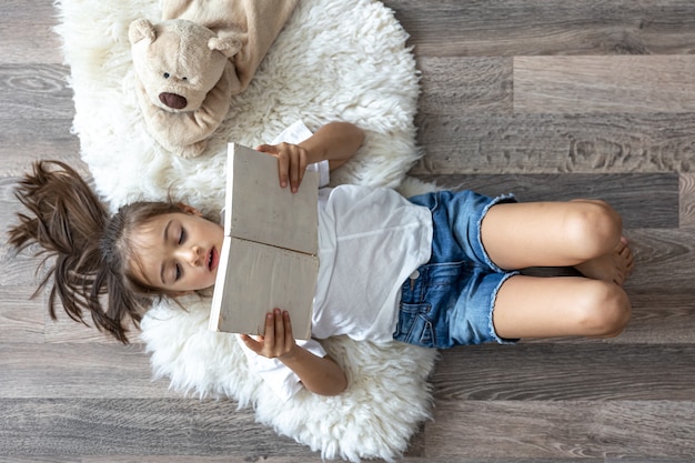 Das Kind liest zu Hause mit seinem Lieblingsspielzeug-Teddybär ein Buch, das auf einem gemütlichen Teppich liegt.