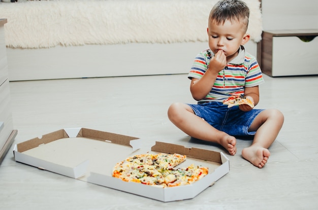 Das Kind sitzt auf dem Boden und isst sehr appetitlich und gierig Pizza, in Shorts und T-Shirt