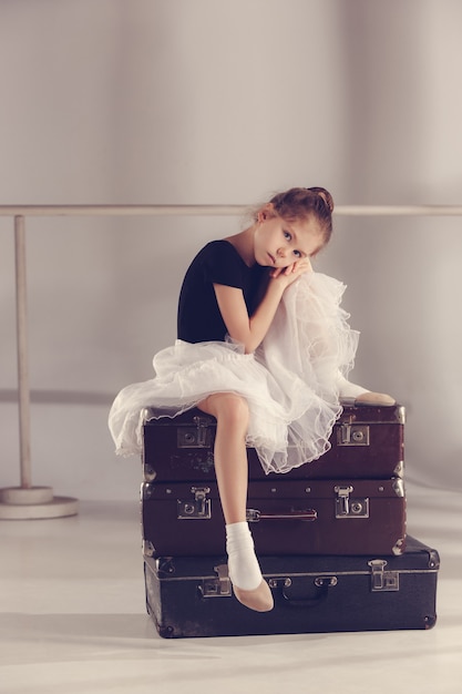 Das kleine Mädchen als Balerina-Tänzerin sitzt im Studio auf Koffern auf grauem Hintergrund. Die Träume von Ballettkonzepten