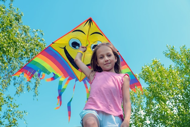 Das kleine Mädchen hält einen hellen Drachen in den Händen und lächelt gegen den blauen Himmel. Konzept von Sommer, Freiheit und glücklicher Kindheit.