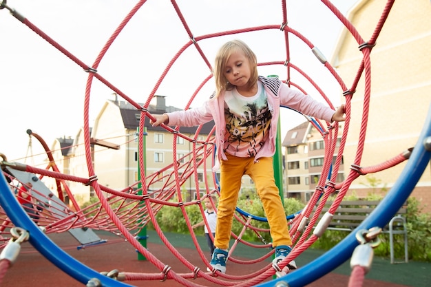 Das kleine Mädchen klettert im Sommer auf dem Spielplatz vor dem Hintergrund der Häuser Seile