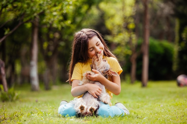 Das kleine Mädchen sitzt mit einem Yorkshire-Terrier auf einem grünen Rasen Der Hund leckt das Mädchen