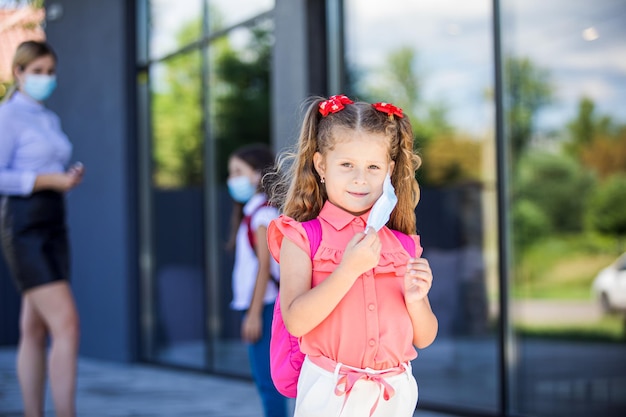 Das kleine Mädchen steht draußen auf einem Schulhintergrund und nimmt eine Schutzmaske ab