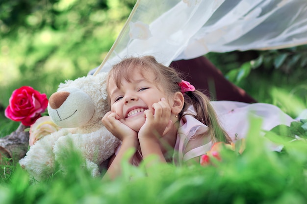 Das kleine Mädchen wirft das Lügen draußen mit einem Teddybären auf und lacht