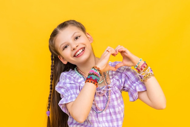 Das kleine Mädchen zeigt ihr Herz mit ihren Fingern und lächelt glücklich Das Kind setzt viele Perlenarmbänder auf Perlen für Kinder Gelber isolierter Hintergrund