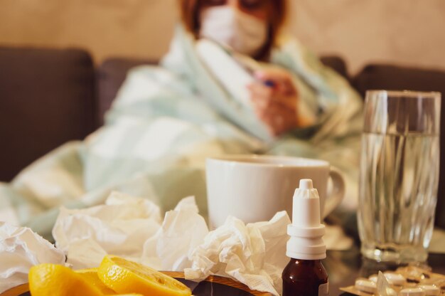 Foto das konzept der erkältung oder saisonalen grippe ein junges mädchen in einer maske schaut auf ein thermometer, das auf einem sofa sitzt, das in eine decke in der nähe eines tisches mit medikamenten gehüllt ist trinkt ein heißes heilgetränk quarantäne