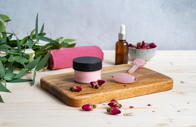 Das Konzept der Gesichts- und Körperpflegekosmetik mit Rose und Eukalyptus Gesichtscreme und Massagegerät auf einem Holztisch mit getrockneten Rosenblüten