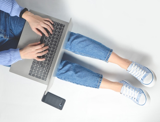 Das Konzept des Bloggens, Arbeiten an einem Laptop. Mit einem Laptop auf einem weißen Boden sitzen. Moderne Technologien. Weibliche Beine in Jeans und Turnschuhen. Draufsicht. Flach liegen.