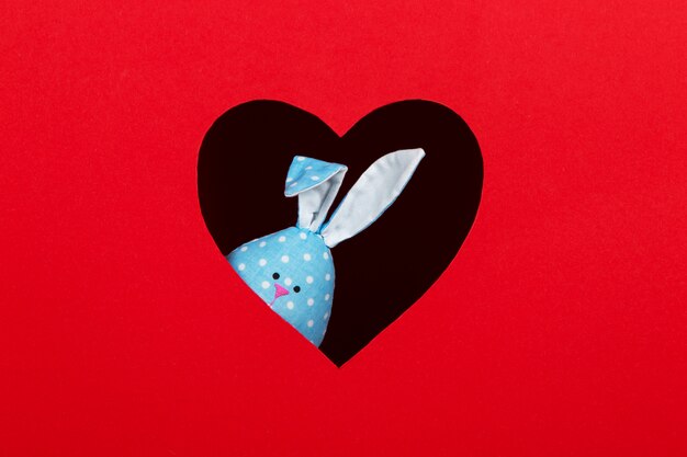 Das Konzept des Frühlings, frohe Ostern, ohrblaues Kaninchen handgemacht, spähen aus einem Loch in der Form eines Herzens auf einem roten Hintergrund. Speicherplatz kopieren.