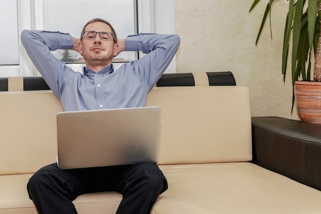 Das Konzept des Ruhebedürfnis während der Arbeit. Ein müder Manager im blauen Hemd streckt sich zufrieden, während er einen Laptop auf dem Schoß hält.