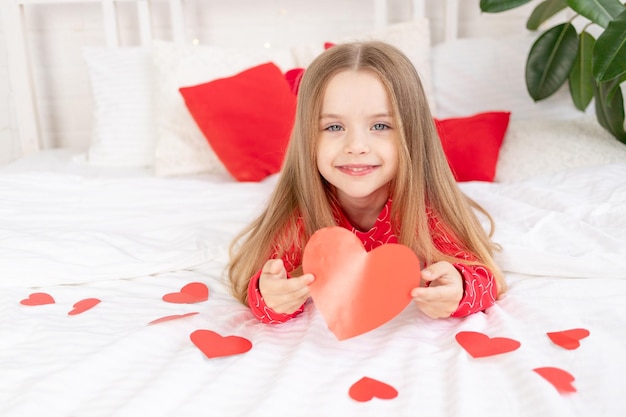 Das Konzept des Valentinstags ein süßes Mädchen auf einem weißen Bett zu Hause in roten Pyjamas zwischen Herzen, die ein Valentinsherz in ihren Händen halten und vor Glück lächeln und zum Urlaub gratulieren