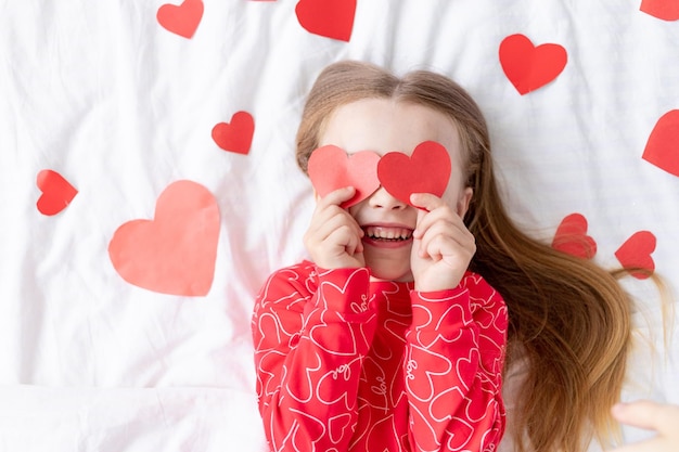 Das Konzept des Valentinstags, ein süßes Mädchen, das zu Hause in einem roten Pyjama zwischen Herzen auf einem weißen Bett liegt, bedeckt ihr Gesicht mit Herzen, verwöhnt sich und täuscht herum und lächelt vor Glück