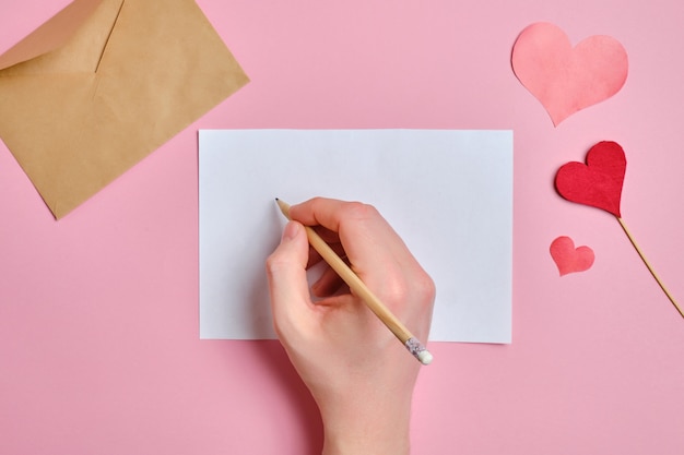 Das Konzept, einen Liebesbrief zu schreiben. Eine Hand hält einen Bleistift über ein weißes Blatt Papier neben Herzen auf einem rosa Hintergrund.