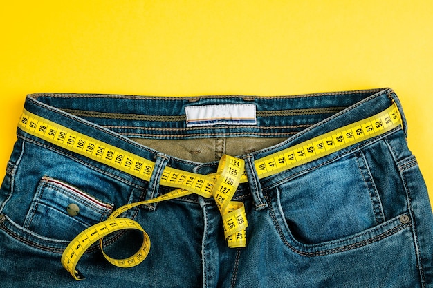 Das Konzept eines gesunden Lebensstils und einer gesunden Ernährung. Blue Jeans mit gelbem Maßband statt Gürtel.