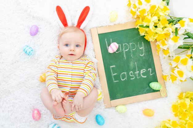 Das Konzept von Ostern ein Baby in einem Hasenhut auf einem Bett mit Ostereiern und der Aufschrift frohe Ostern