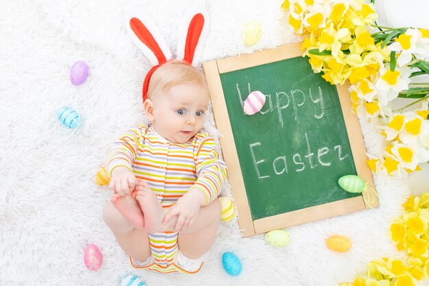 Das Konzept von Ostern, ein Baby in einem Hasenhut auf einem Bett mit Ostereiern und der Inschrift frohe Ostern