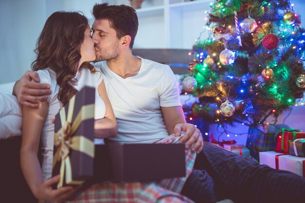 Das küssende Paar packt die Geschenkbox neben dem Weihnachtsbaum aus. Abendzeit