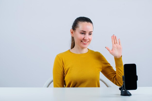 Das lächelnde Blogegr-Mädchen zeigt eine Hallo-Geste, indem es vor einer mobilen Kamera auf weißem Hintergrund sitzt