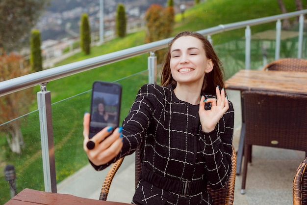 Das lächelnde Blogger-Mädchen macht ein Selfie mit der Rückkamera des Smartphones, indem es eine Okay-Geste auf dem Hintergrund der Naturansicht zeigt