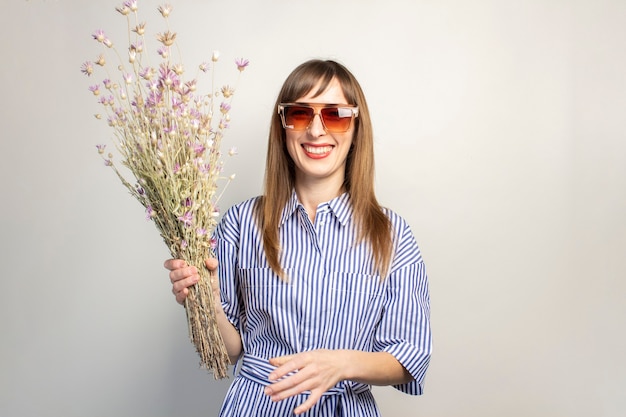 Das lächelnde junge Mädchen in der Sonnenbrille hält einen Strauß Wildblumen auf einem hellen Hintergrund