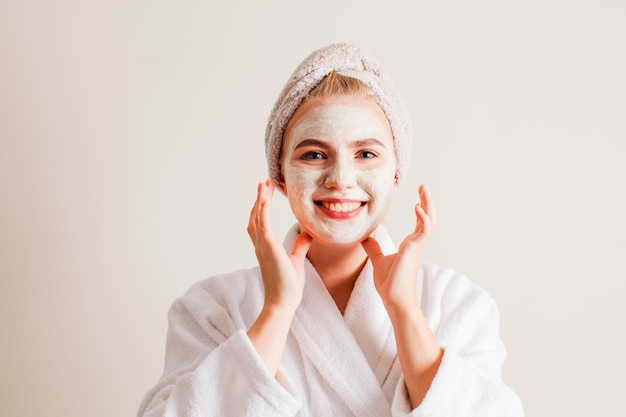 Das lächelnde schöne Mädchen mit cremefarbener Gesichtsmaske steht in einem Bademantel mit einem Handtuch auf dem Kopf vor dem Hintergrund der Wand