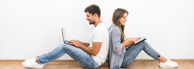 Das Lächelnpärchen sitzt mit einem Laptop und einem Tablet in der Nähe der leeren Wand