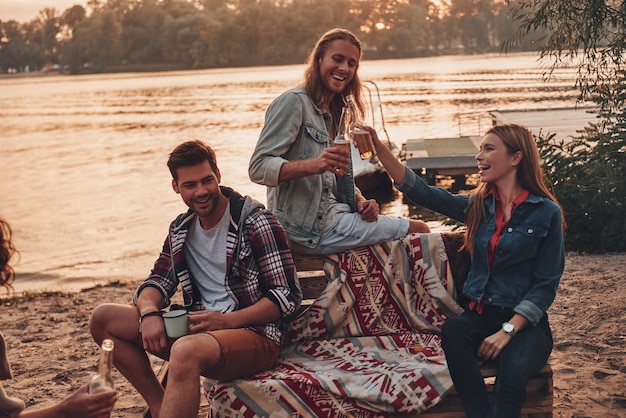 Das Leben ist großartig! Gruppe junger Leute in Freizeitkleidung, die sich lächeln und anstoßen, während sie eine Strandparty in der Nähe des Sees genießen?