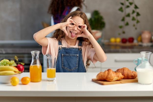 Das lustige kleine Mädchen mit dem lockigen Haar, das Grimase macht, steht zusammen mit der Mutter in der Küche.