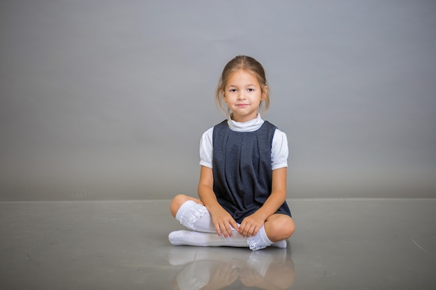 Das Mädchen als Erstklässlerin in Schuluniform - ein graues Sommerkleid und eine weiße Bluse posieren auf Grau