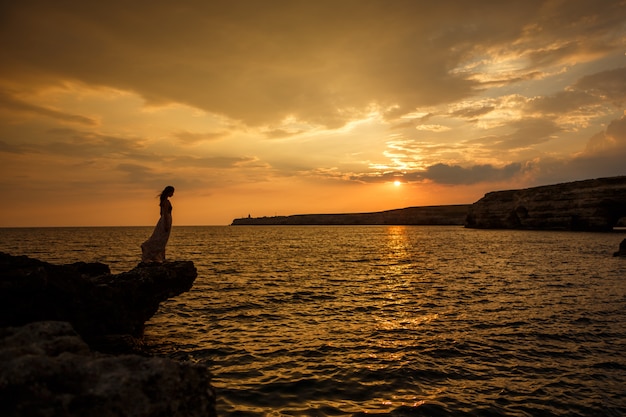 Das Mädchen auf dem Hintergrund eines schönen Meerblicks und des Sonnenuntergangs, Schattenbild eines Mädchens auf einer Klippe, auf einer Klippe, einem schönen Himmel und einem Meer