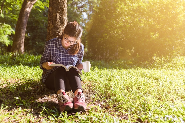 Das Mädchen, das auf einem grünen Gras mit Tasse Kaffee sitzt und liest das Buch