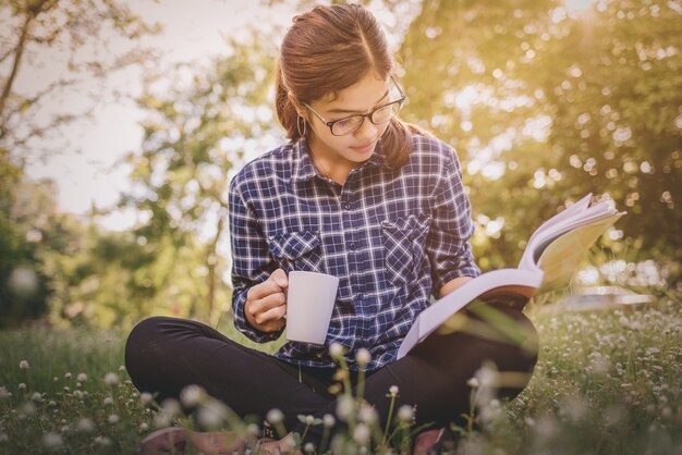 Das Mädchen, das auf einem grünen Gras mit Tasse Kaffee sitzt und liest das Buch