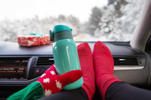 Das Mädchen hält eine Thermoskanne, während es im Winter im Auto auf dem Hintergrund des Winterwaldes sitzt Reisereise und Winterkonzept