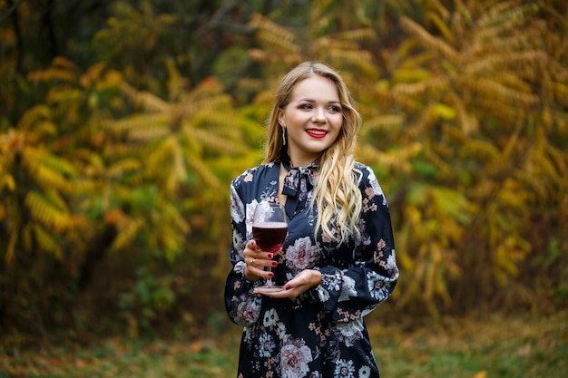 Das Mädchen im Kleid hält ein Glas Rotwein im Herbstwald