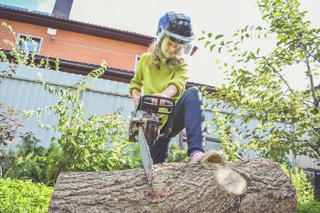 Das Mädchen in einem Helm mit einer elektrischen Säge sägt einen Baumstamm