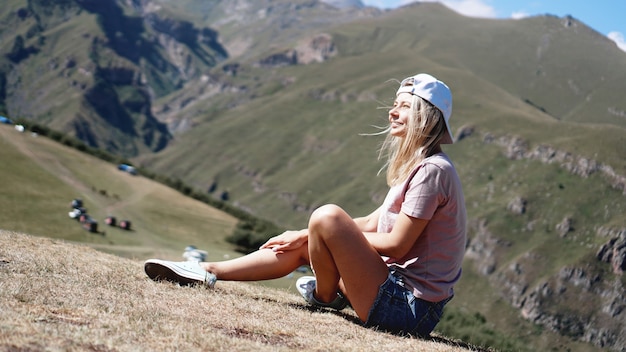 Das Mädchen in Mütze und Shorts in den Bergen sitzt auf dem Gras