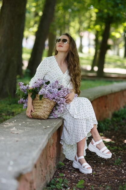 Das Mädchen in weißem Kleid und Sonnenbrille hält in ihren Händen einen Weidenkorb mit Blumen. Korb mit Flieder. Mädchen und Blumen. Setzen Sie sich mit einem Korb voller Flieder in die Hände. Floristik