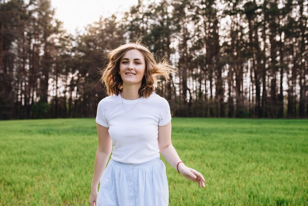 Das Mädchen ist blond, braunes Haar, trägt ein weißes Hemd und einen blauen Midirock. Auf dem Feld durch das grüne Gras gehen. Porträt eines Mädchens. Positiv und lächeln.