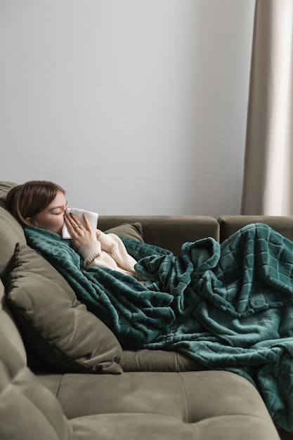 Das Mädchen ist krank und wischt sich mit einer Papierserviette die Nase ab, während es in eine Tagesdecke gehüllt auf der Couch liegt