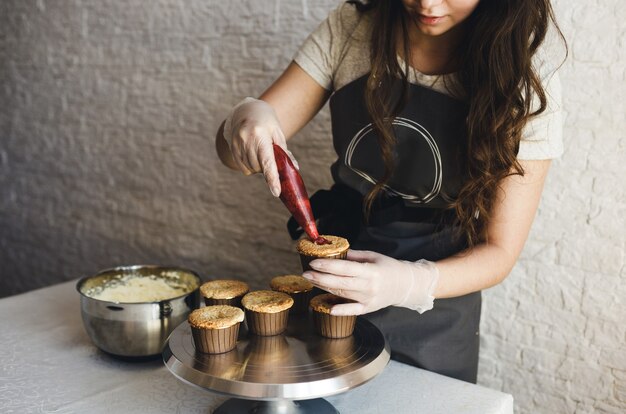 Das Mädchen kocht zu Hause Cupcakes und füllt sie mit Füllung. Hände eines Küchenchefs mit einem Spritzbeutel, der die Sahne auf die Cupcakes drückt.