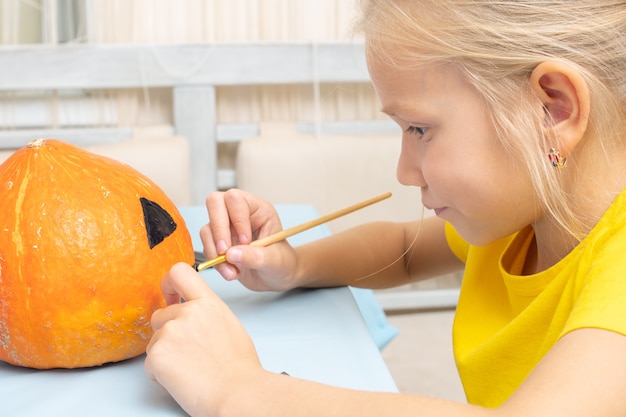 Das Mädchen kreiert eine Laterne aus einem Kürbis und sitzt zu Hause am Tisch in der Küche. Halloween-Party und Familienlebensstil-Hintergrund