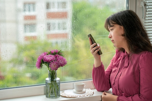 das mädchen schreibt sms auf einem handy, das neben einem regnerischen fenster steht.