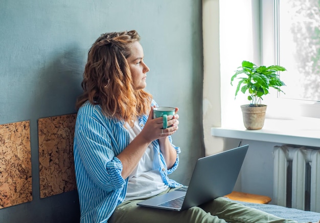 das mädchen sitzt auf dem bett am computer mit einer tasse kaffee tee freiberufliche arbeit zu hause eine fernbedienung