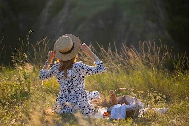 Das Mädchen sitzt im Gras in der Natur. Das Mädchen schaut auf den Sonnenuntergang. Das Mädchen ruht in der Natur mit einem Korb mit Essen. Picknick in der Natur.